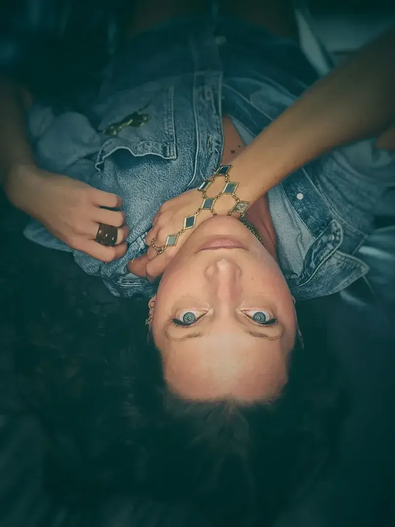 Elle s’était allongée là, sans en connaître la raison exacte. Peut-être pour échapper au vacarme du monde, ou pour se perdre enfin dans le murmure d’elle-même — nue d’artifice, déliée de toute attente.La lumière effleurait sa peau comme une confidence glissée à l’aube, trop intime pour être dite, trop tardive pour être tue. Sur le velours pâle de son épaule, le jour s’attardait, hésitant, tremblant d’envie.Le denim froissé gardait l’empreinte de souvenirs indociles : un été suspendu, un souffle retenu, un regard croisé dans un miroir fissuré. Autour de son poignet, les bijoux frémissaient, capturant la clarté pour la rendre au monde en mille éclats d’or — éclats d’elle-même, éparpillés, insaisissables.Chaque reflet la dédoublait, la recomposait autrement.L’une riait encore, bouche entrouverte sur un secret.L’autre pleurait d’avoir trop aimé.Une troisième, plus sauvage, guettait l’ombre du désir — prête à s’y dissoudre.Elle inspira, lentement.Le silence vibrait, comme une peau.Et dans le miroitement trouble de ses pensées, elle reconnut enfin celle qu’elle avait laissée derrière les masques : la femme entre les reflets.Un sourire effleura sa bouche, à peine un souffle.Les fragments ne blessaient plus.Ils brûlaient, d’une lumière douce et dangereuse.